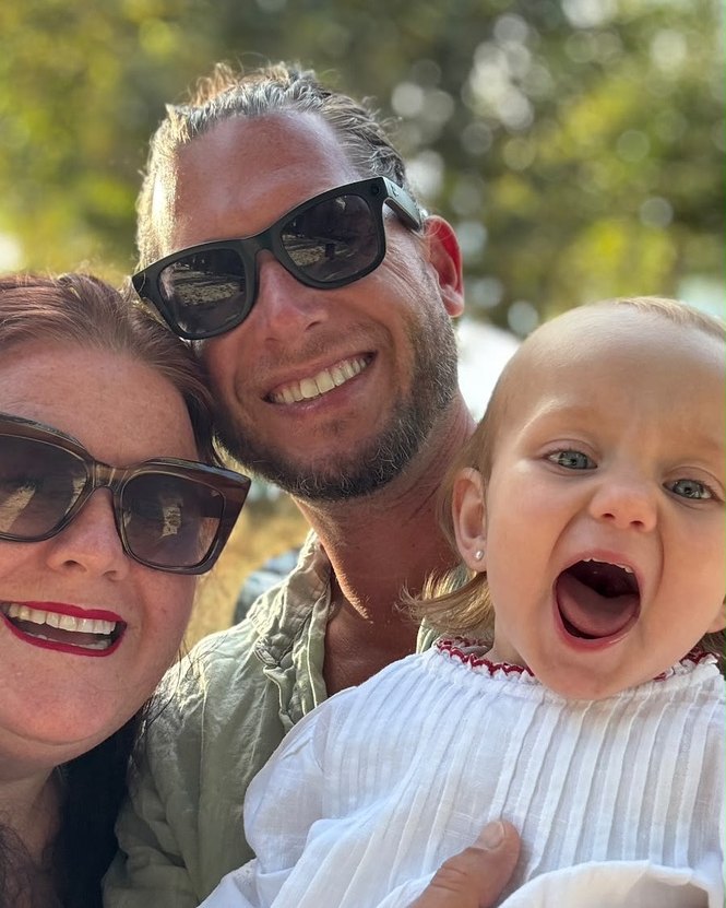 Andy and Anja Gibson smiling close together outdoors, holding their baby daughter, who is laughing with her mouth wide open.