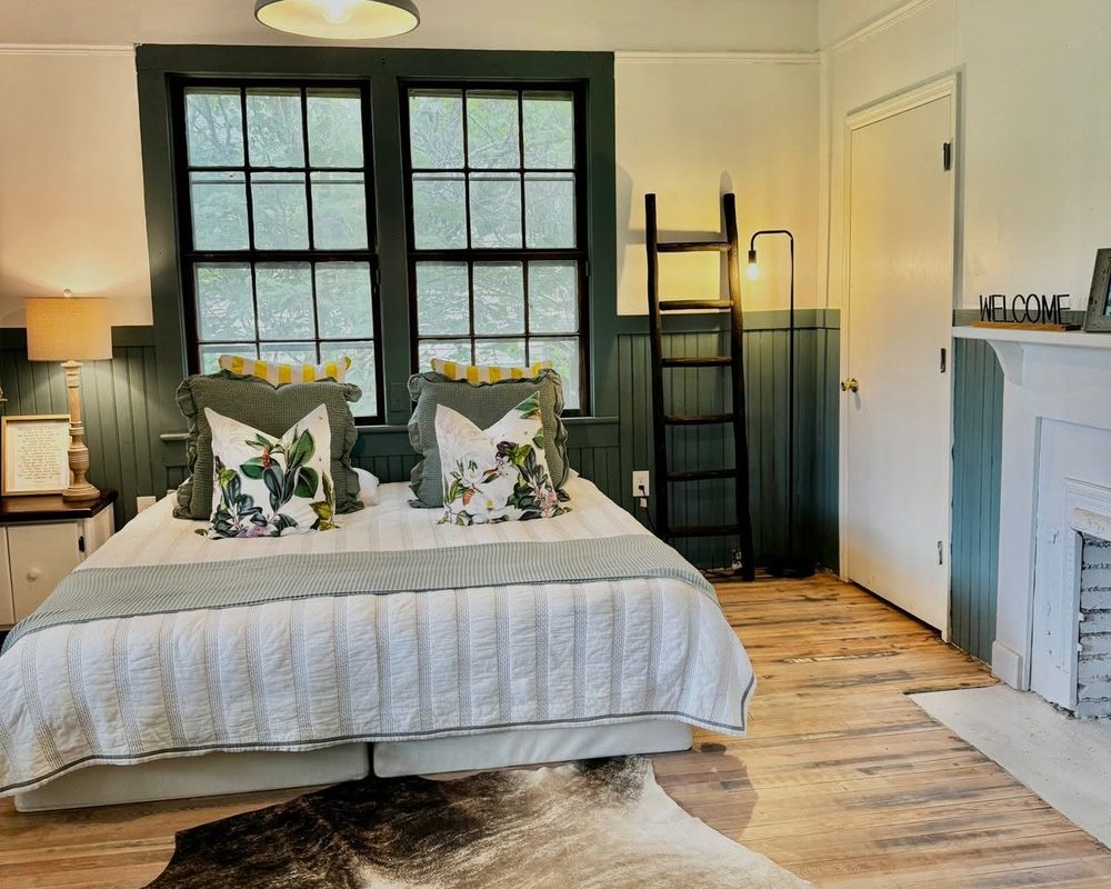 The same bedroom after restoration: painted green wainscoting, tall windows, a made bed, a wooden ladder, and refinished original floors.