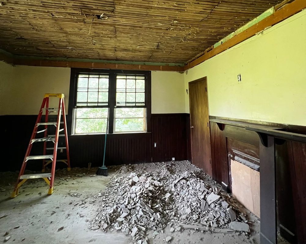 A bedroom at 403 I Street before restoration: bare lath ceiling, dark wainscoting, debris on the floor, and a ladder by the windows.