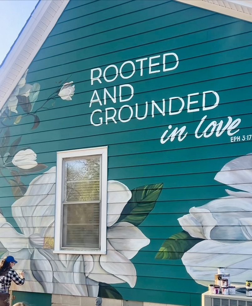 A large mural on the side of a restored mill house reading “Rooted and grounded in love, Ephesians 3:17,” with white magnolia blossoms on a deep teal wall.